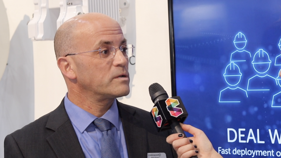 A person with glasses, a bald head, and dressed in a blue suit and tie is speaking into a microphone with a colorful logo. They are standing beside a screen displaying blue graphics of people and the partial text "DEAL WITH" and "Fast deployment..." in a professional setting.