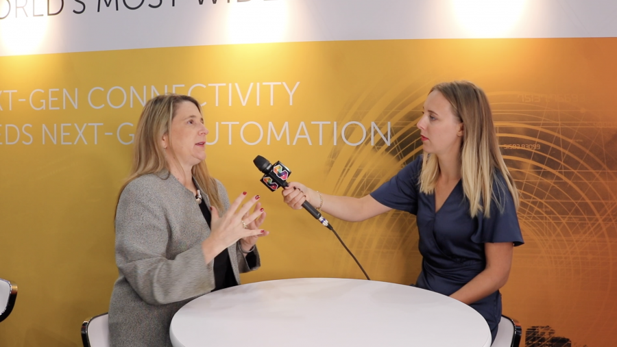 Two women are seated at a round white table. The woman on the left is gesturing with her hands, wearing a gray blazer. The woman on the right, holding a black microphone, wears a navy blue dress. Behind them is a yellow and white backdrop with text about connectivity and automation.