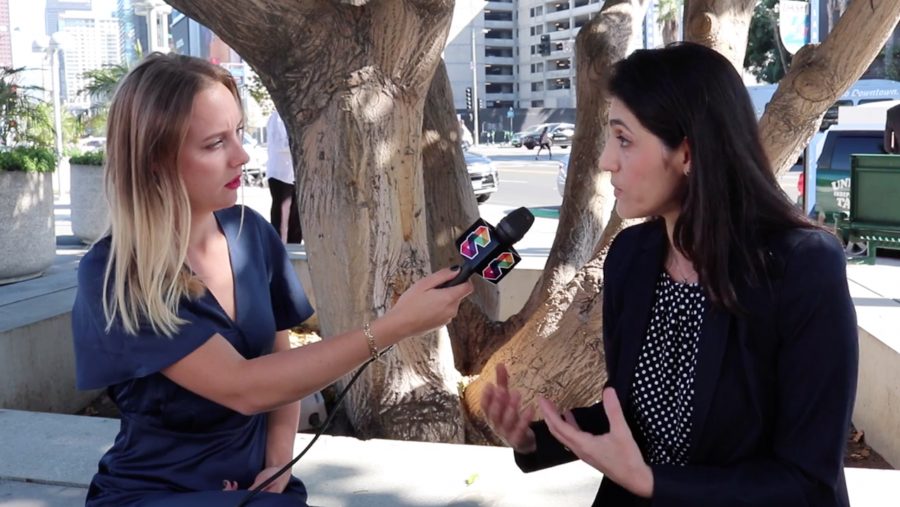Two women are seated outdoors in front of a large tree. The woman on the left, in a blue dress, holds a colorful microphone towards the woman in a dark blazer on the right. They appear to be engaged in an interview. Urban buildings and a street scene are visible in the background.