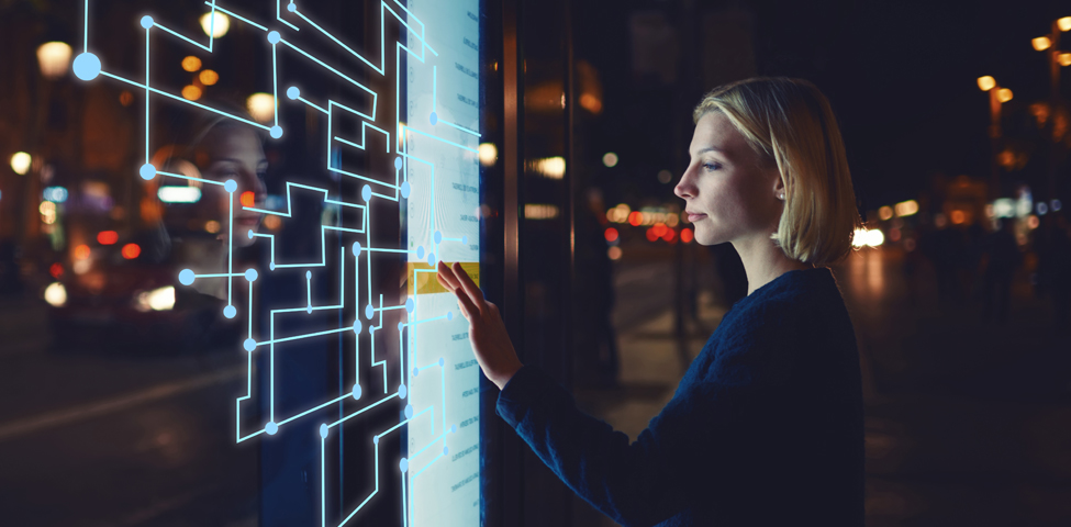 A woman interacts with a glowing touchscreen in a nighttime urban setting. The display shows a network of connected lines and nodes. She appears focused, with city lights blurred in the background, creating a futuristic ambiance.