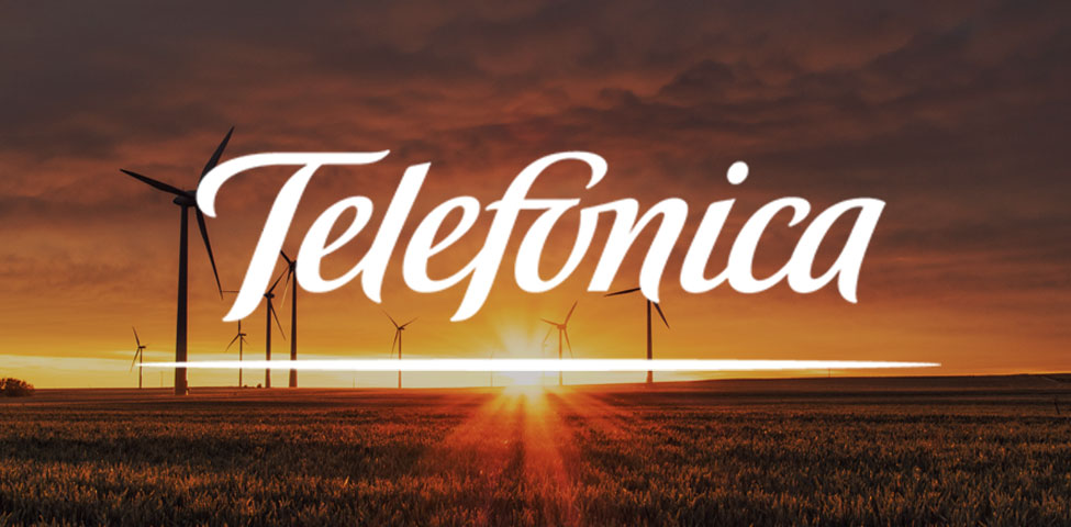The image shows a sunset over a field with wind turbines silhouetted against the sky. The word "Telefonica" is prominently displayed in stylized white text across the center. The sky is a mix of orange and dark hues, adding a warm ambiance to the scene.