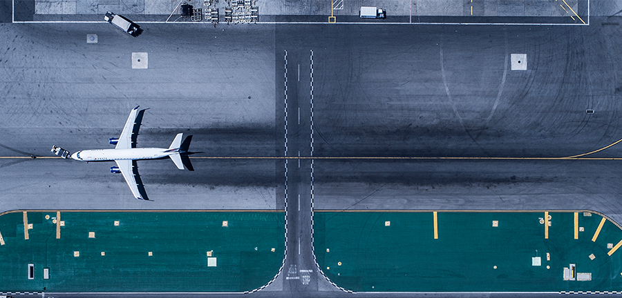 Aerial view of an airport runway featuring a stationary airplane positioned on the left side. The runway displays distinct markings and a green section at the bottom, possibly indicating a taxiway. Various equipment and vehicles are visible near the top of the image, highlighting airport operations.