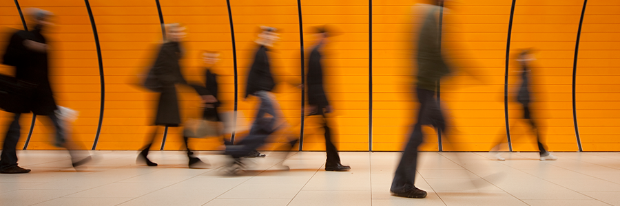 Blurred image of people walking past an orange, curved-panel wall in a subway station. The scene conveys motion, with figures in dark clothing moving horizontally from left to right. The wall panels create a repetitive background pattern against the light-colored floor.