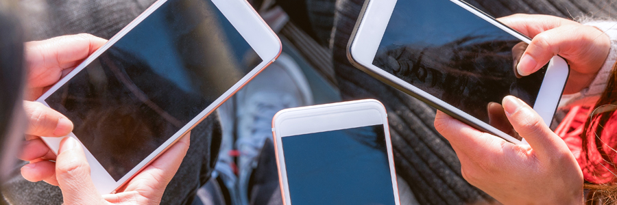 Four hands holding smartphones with blank screens in a close circle, seen from above. The phones have white and black cases. The background is blurred, suggesting an outdoor setting. The people are wearing casual clothing, indicating a relaxed, social gathering.