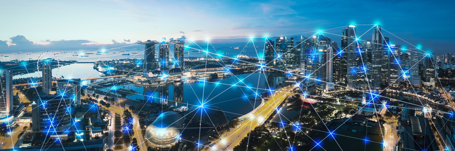A cityscape at dusk with illuminated skyscrapers and buildings. The foreground features a river with bridges. Blue network-like lines and glowing nodes overlay the scene, symbolizing connectivity and technology. The sky transitions from blue to orange as the sun sets.