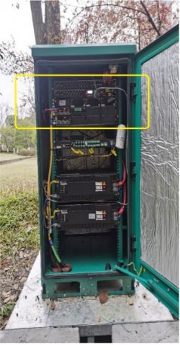The image shows an open green outdoor electrical cabinet containing various electronic components and wires. The inner door is lined with silver insulation. Three large black units with visible labels are stacked vertically, connected by red and green wires. A yellow rectangle highlights the top section.