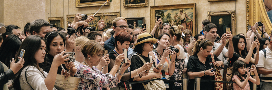 A diverse crowd of people are gathered in a museum, capturing photos with smartphones and cameras. Some hold up selfie sticks, while others focus on specific objects or artwork. Paintings are visible on beige walls, and the atmosphere is lively and engaged.