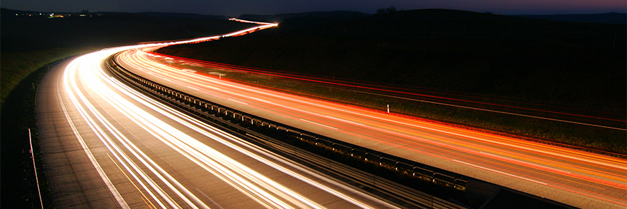 Long exposure photo of a highway at night showing bright white and red light trails. The white trails are on the left, indicating vehicles moving away, while red trails are on the right, indicating vehicles approaching. The surrounding landscape is dimly lit under a dark sky.