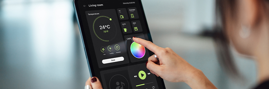 A person adjusts smart home settings on a tablet. The screen shows features like temperature control set at 24°C, lighting options with a color wheel, and music playback controls. The background is blurred, emphasizing the focus on the device and hand interaction.