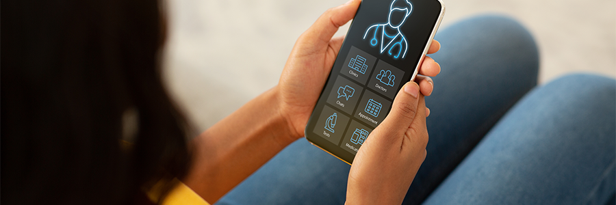 A person is holding a smartphone displaying a health app. The screen shows icons for appointments, patient care, chat, calendar, and prescriptions. The person wears a yellow shirt and blue jeans, sitting with their knees bent, viewing the device closely.