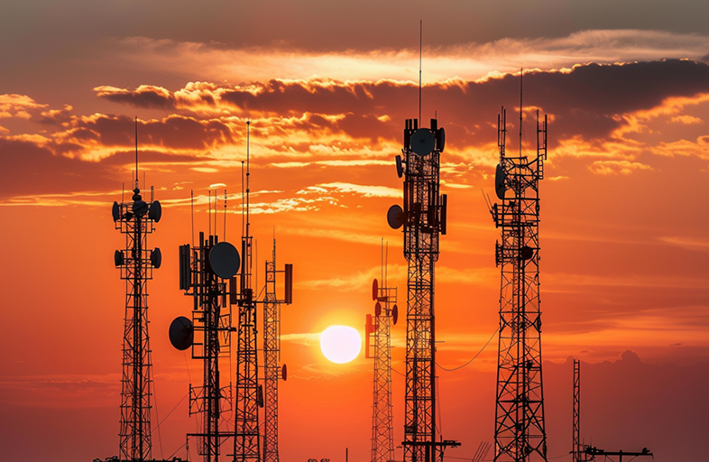 Silhouetted against a vibrant orange and red sunset sky, several communication towers stand tall with antennas and dishes visible. The sun sets behind the towers, casting a warm glow and highlighting the clouds in the background.