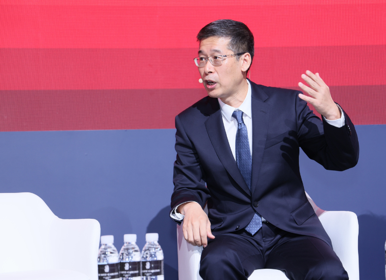 A man in a suit sits on a white chair, speaking energetically with his right hand raised. He wears glasses, a dark suit, and a tie. A row of four water bottles is on the table beside him. The background features red and blue horizontal stripes.