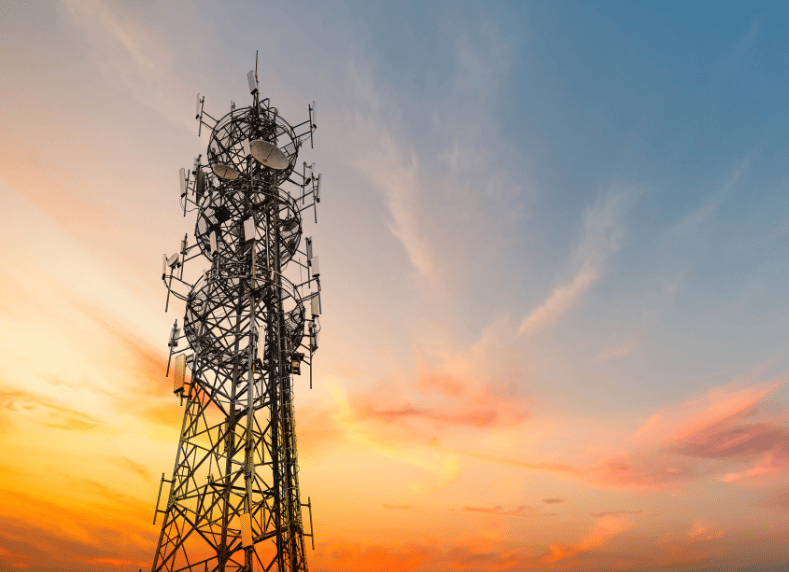 A tall communication tower stands against a vibrant sunset sky with various antennas and satellite dishes attached. The sky is a blend of orange, pink, and blue hues, creating a striking backdrop. The intricate metal framework of the tower is silhouetted against the colorful sky.