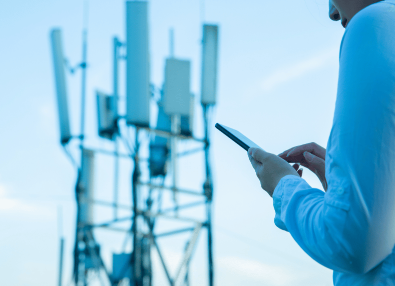 A person in a white shirt holds a smartphone, standing outdoors. In the background, a large cell phone tower with multiple antennas is visible against a clear blue sky. The image suggests communication or technology themes.