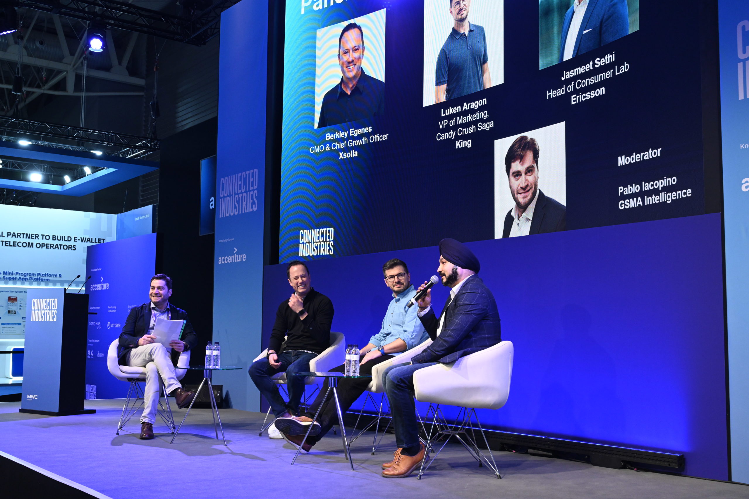 A panel of five men seated on a stage under the "Connected Industries" banner, engaged in discussion. Each has a microphone. A large screen behind them displays their names and titles. An audience sits in a dimly lit area. The event appears professional and focused on telecom operators.