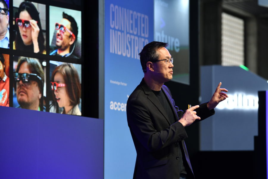 A speaker in a black suit gestures on stage at a tech conference. A large screen displays images of people wearing augmented reality glasses. The stage backdrop reads “Connected Industries” with partner logos. The scene suggests a presentation on technology innovations.