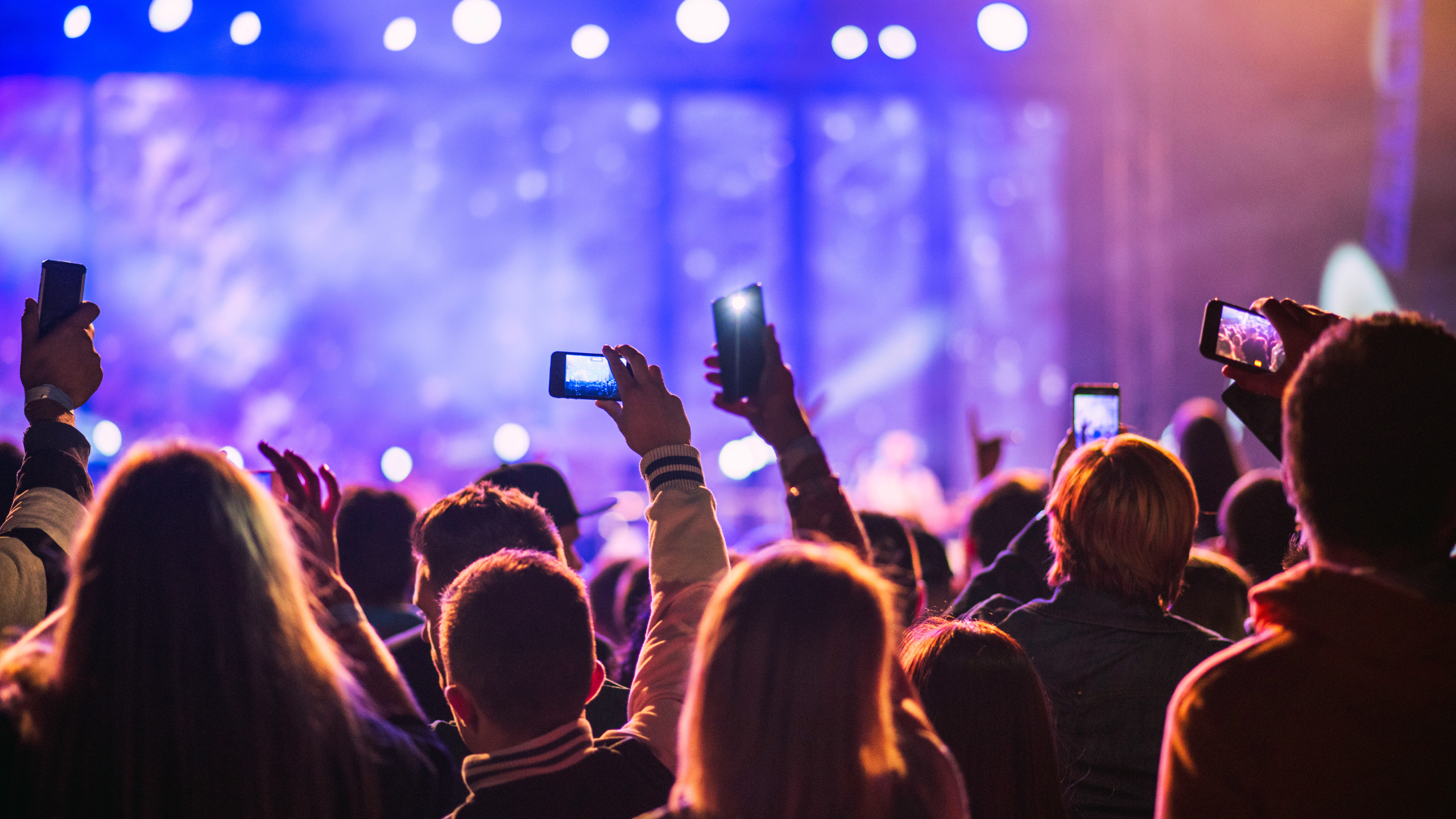A lively concert scene with a crowd of people facing a brightly lit stage. Many attendees hold up smartphones to record the performance. Colorful lights cast a vibrant glow over the audience. The atmosphere is energetic and filled with anticipation.