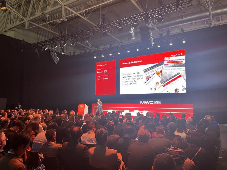 A speaker stands on a stage in front of a large audience. Behind them is a screen displaying a presentation slide titled "Problem Statement," with text and images. The event is in a dimly lit room, featuring modern lighting and a high ceiling. Audience members are attentively watching.