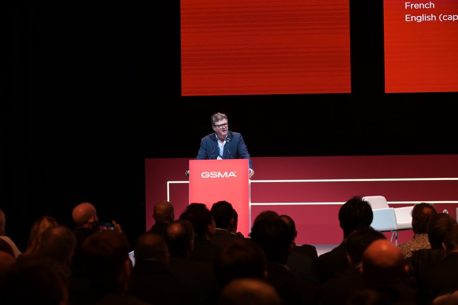 A person in a suit speaks at a podium labeled "GSMA" on a stage with a red background. They are addressing an audience in a dimly lit setting. Two screens above them display text in various languages. An empty chair is positioned to the right of the podium.