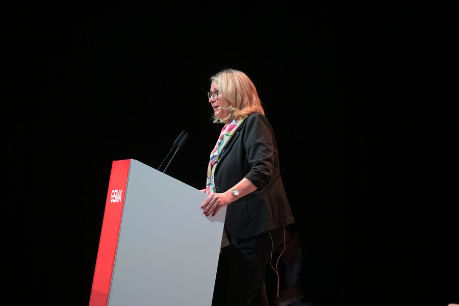 A woman with blonde hair wearing glasses is speaking at a podium on a stage. She is dressed in a black suit with a multicolored scarf. The podium is red and gray with "GSW" written on it. The background is dark, highlighting the speaker under the stage lights.