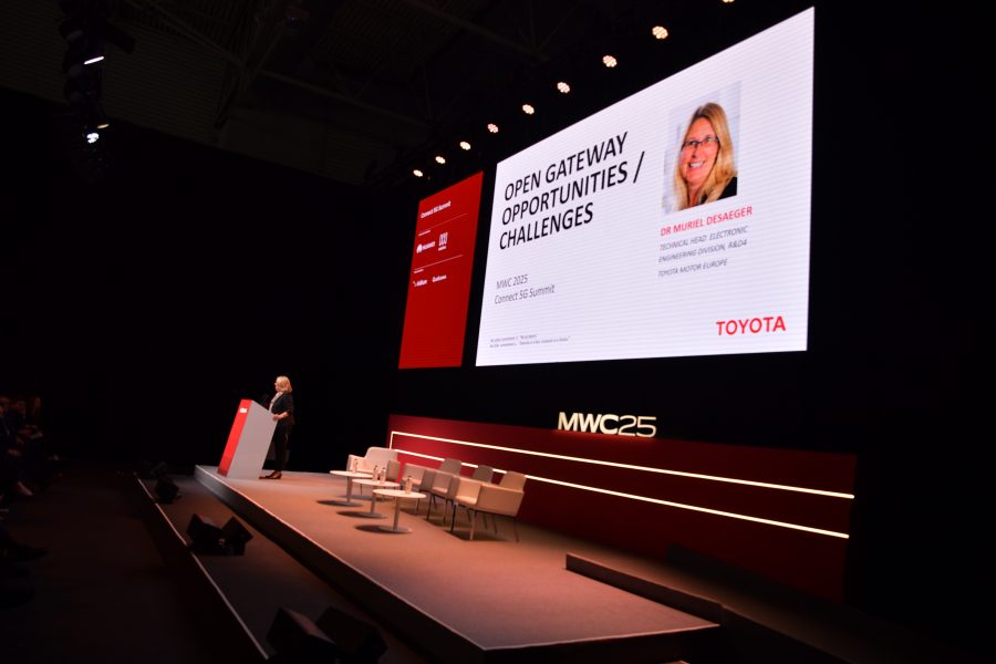 A woman stands at a podium on a stage, addressing an audience. A large screen behind her displays "Open Gateway Opportunities/Challenges" and a photo of her with details about her role at Toyota. The stage is set with several empty chairs. The event is labeled MWC 2023.