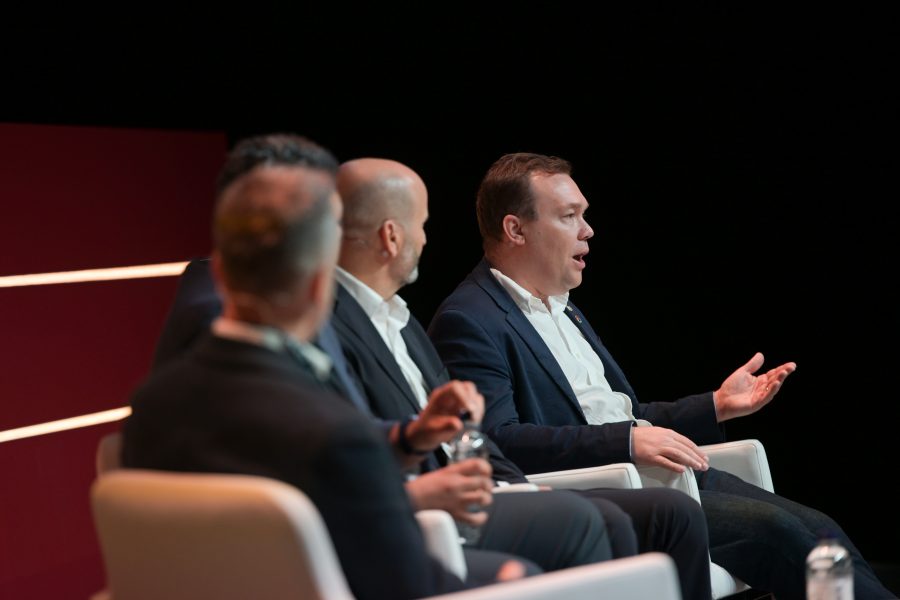 Four men in suits are seated on stage in white chairs, engaged in a discussion. The man in the foreground gestures with his hand while speaking. A dark background contrasts with red horizontal lines on the left. A bottle of water is on the table in front.