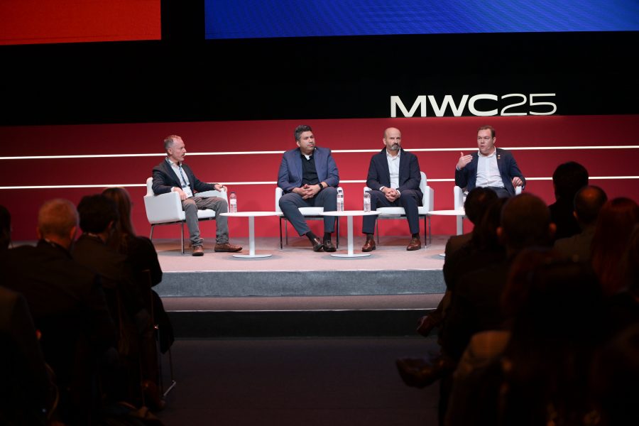 Four men in suits sit on a panel stage at MWC25, with a red and blue backdrop. The audience is visible in silhouette. The speaker on the right is gesturing with his hand, while the others listen attentively. A modern, professional setting focused on discussion and presentation.