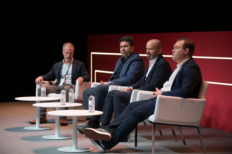 Four men sit on a stage, each in a white chair, participating in a panel discussion. They are wearing suits with open-collared shirts. Three round tables in front hold bottled water. The backdrop is dark red with horizontal white lines, and the stage floor is gray.