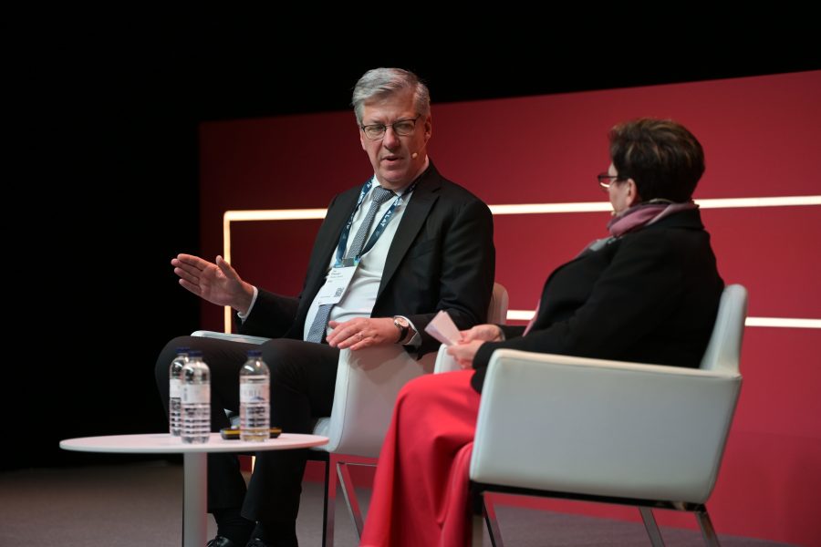 A man and a woman are seated on stage in conversation. The man gestures with his hand, wearing a dark suit and lanyard. The woman, facing him, wears a dark outfit with a red skirt. Two water bottles sit on a round table between them. A red background with a white stripe is behind them.