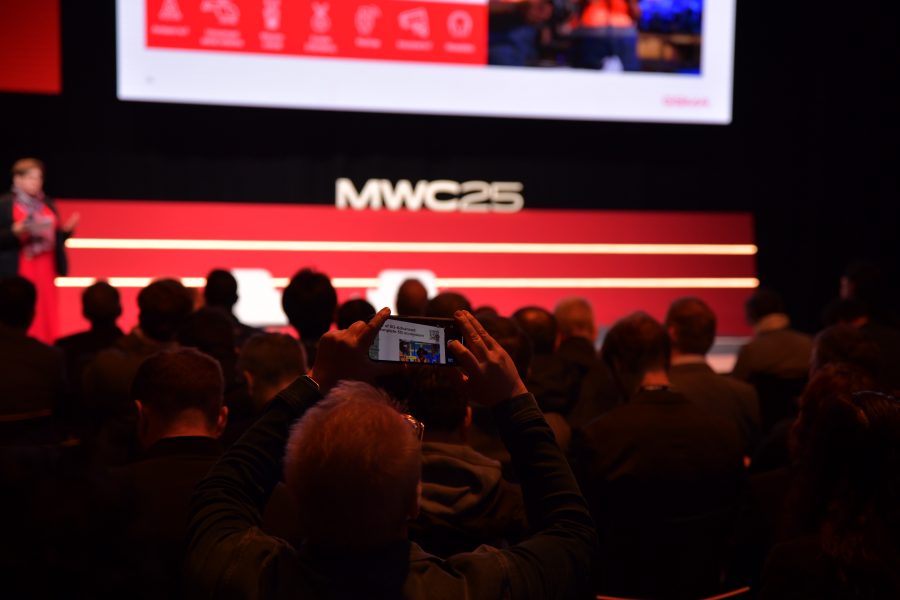 Audience members face a stage at an event marked "MWC25." A person in the foreground captures the presentation on their smartphone. The stage features a large screen displaying colorful graphics. The atmosphere is focused and attentive.