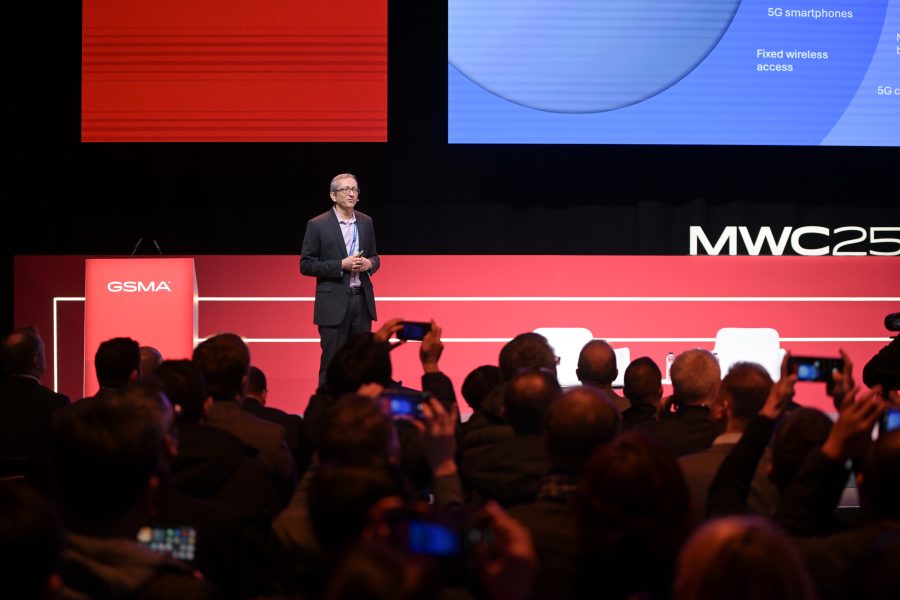 A speaker stands on stage in front of a seated audience at a conference. The backdrop features the logos "MWC25" and "GSMA." Attendees are capturing the presentation with their phones. A large screen behind the speaker displays part of a chart related to 5G technology.