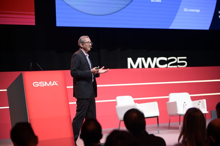 A man in a suit and glasses gestures while speaking on stage at MWC25. He's standing beside a red podium with "GSMA" on it. Behind him, a screen displays a presentation, and two empty white chairs are visible. The audience watches attentively in the foreground.