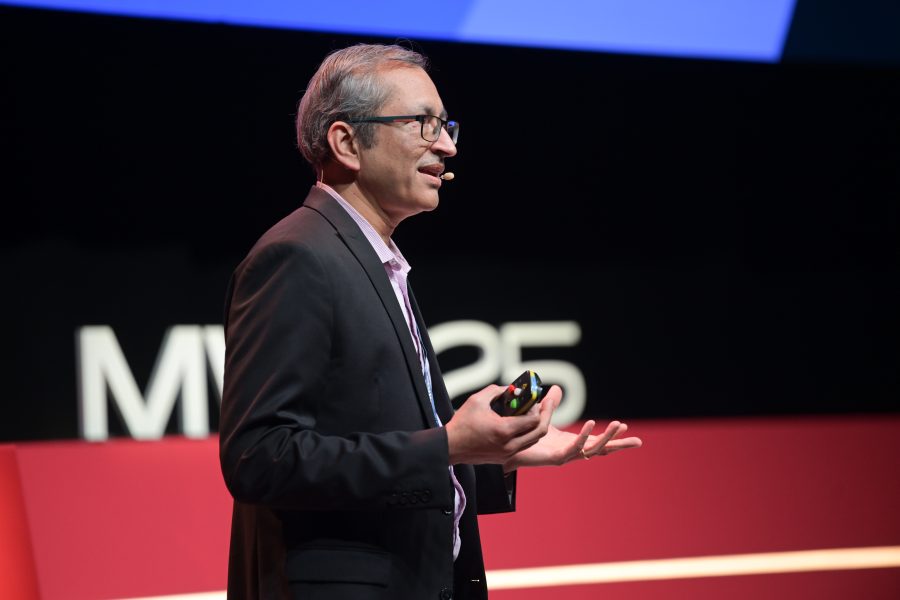 A man wearing a suit and a headset microphone gestures while speaking on stage. He holds a clicker in one hand. Behind him are large letters "MWC23" on a red and black backdrop. The scene suggests a conference or presentation setting.