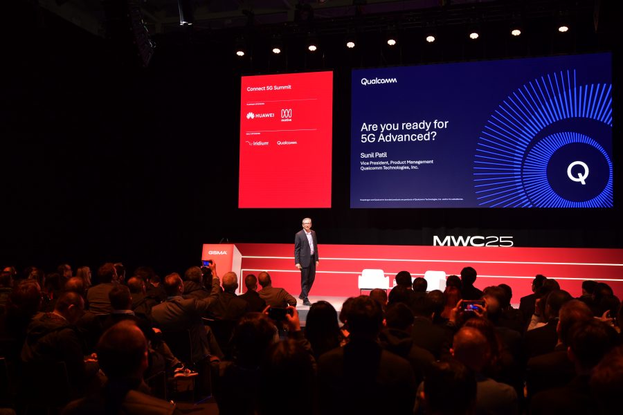 Man standing on stage speaking at a tech conference. Large screen displays "Are you ready for 5G Advanced?" and "Qualcomm." The backdrop is lit with blue and red. Audience seated in foreground. Event labeled "MWC23." Red podium with small screen showing logos of Nokia, Huawei, and others.