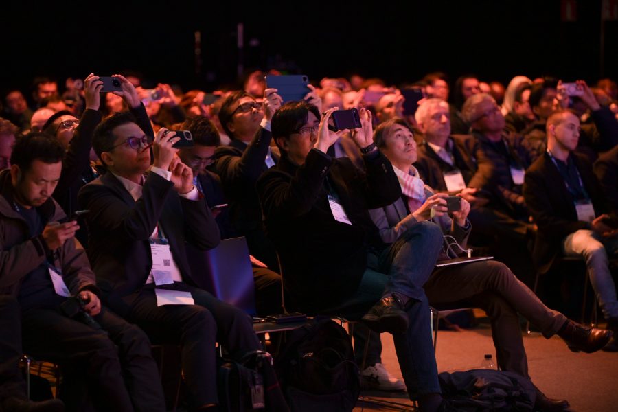 Audience members in a dimly lit venue take photos and videos with smartphones. They are seated closely together, focusing intently on something off-camera to the right. Many are wearing name tags, and the atmosphere suggests a formal event or presentation.