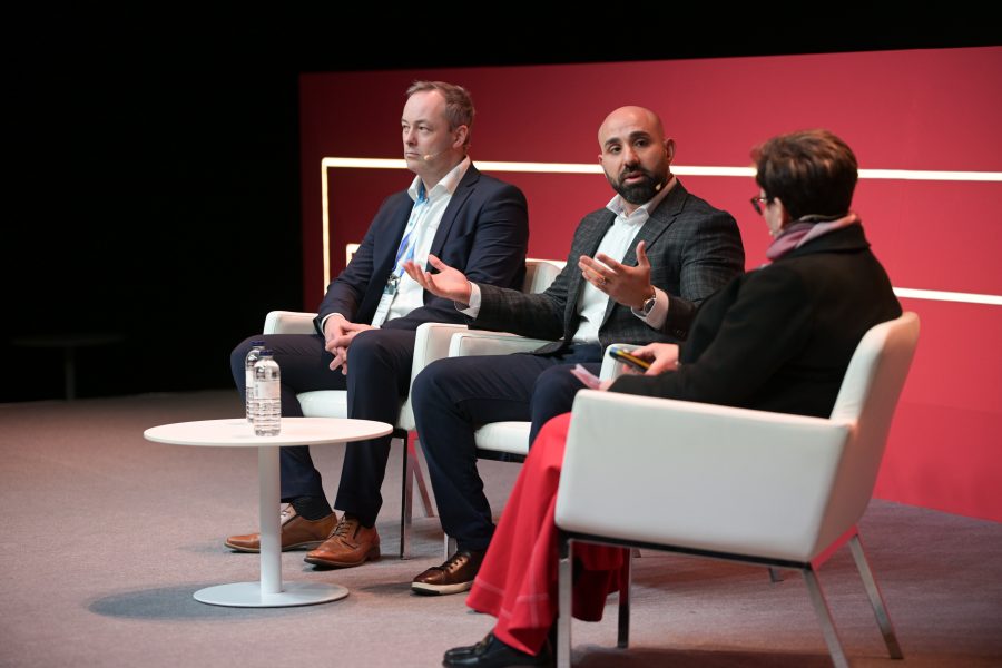Three people seated on stage in a panel discussion. Two men in suits, one speaking and gesturing with hands, the other listening. A woman with short hair and glasses writes notes. White chairs and round table with a water bottle. Red and black backdrop with horizontal lines.