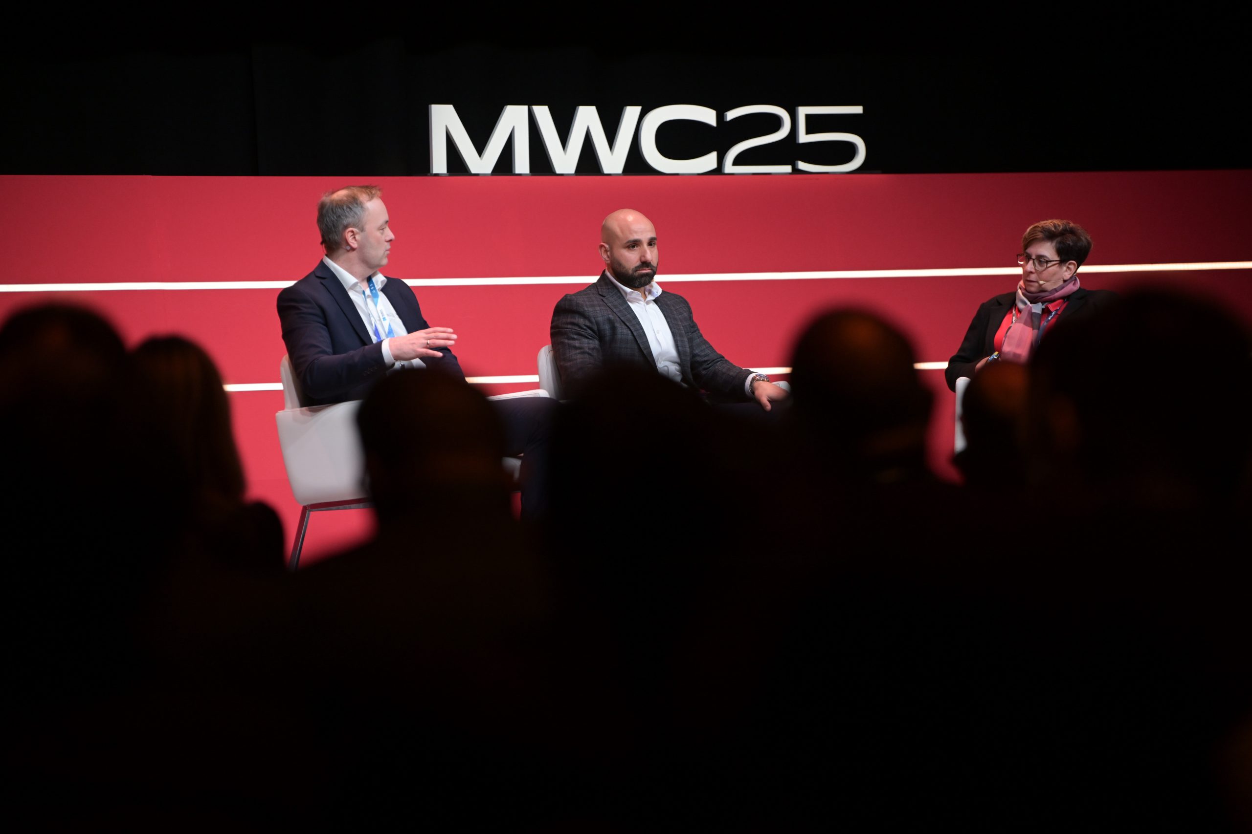 Three individuals sit on stage at MWC25. The person on the left gestures while speaking, the person in the middle listens attentively, and the person on the right looks engaged. They are seated against a red backdrop with the event name "MWC25" displayed prominently. Audience silhouettes are in the foreground.