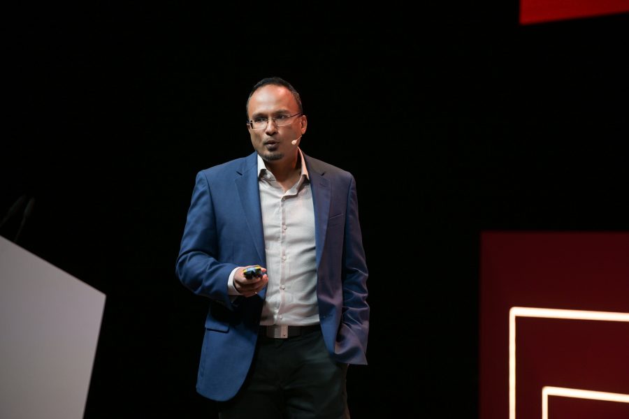 A man wearing glasses, a blue blazer, and a light shirt is speaking on stage. He holds a device in his right hand and has his left hand in his pocket. The background is dark with red geometric shapes. An angled podium is partially visible on the left.