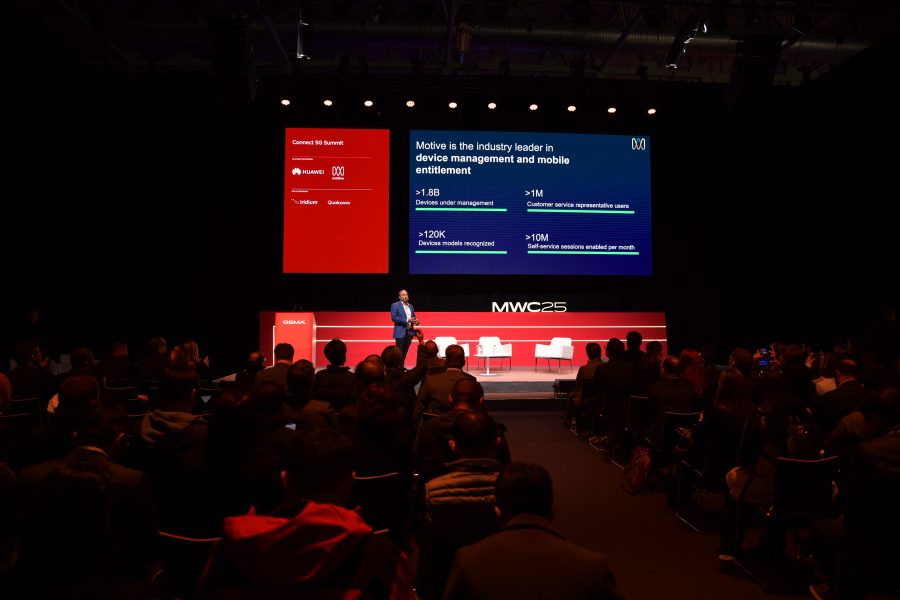 A speaker on stage at MWC 25, delivering a presentation. A large screen displays statistics about device management and mobile entitlement. The audience is seated in a dimly lit auditorium, attentively listening. The stage features red and blue branding elements.