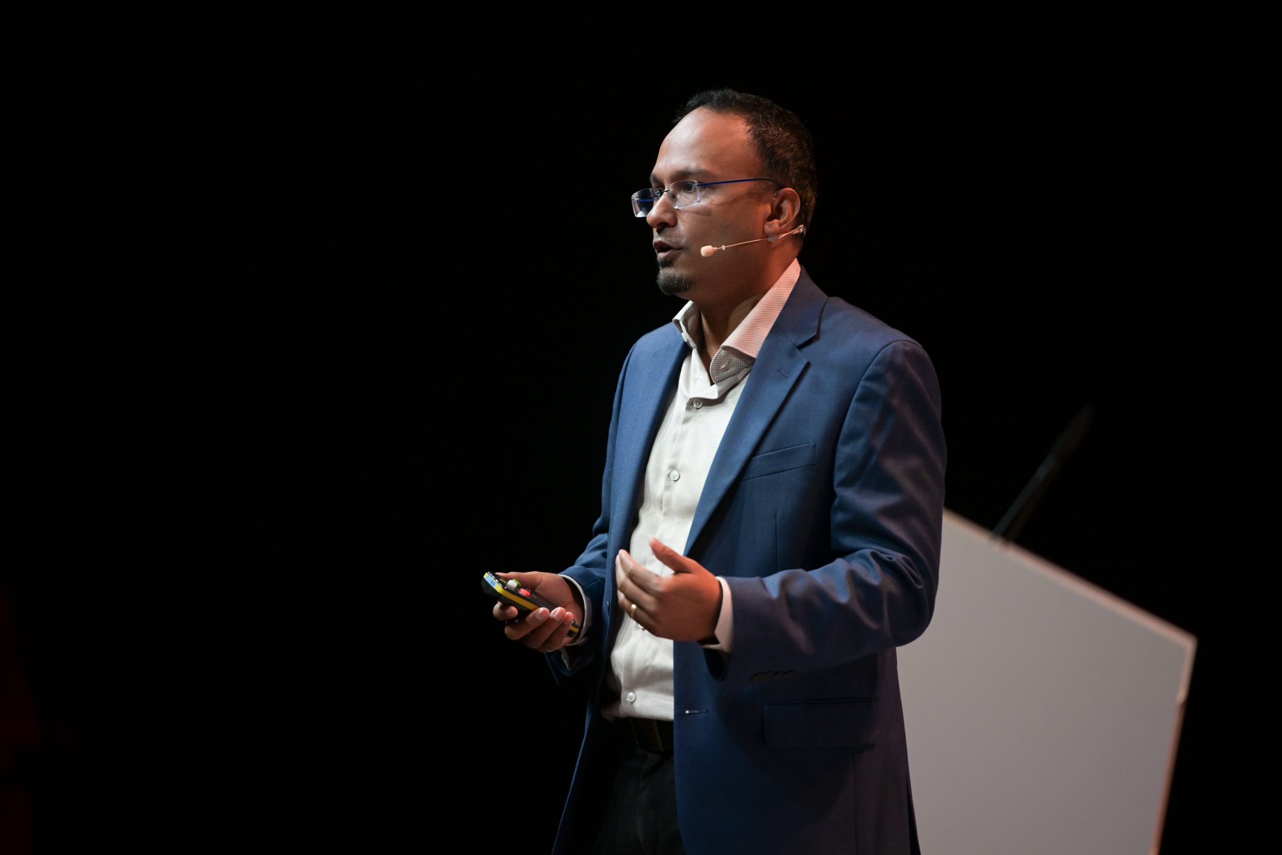 A person in a blue blazer and white shirt speaks on stage with a microphone headset. They hold a device in one hand. The background is dark with a white lectern partially visible, suggesting a presentation or lecture setting.