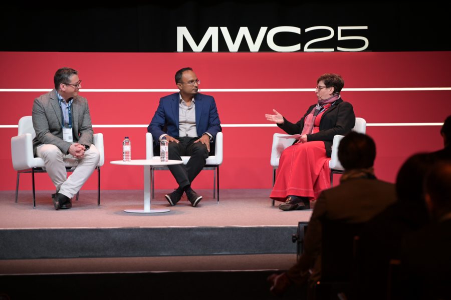 Three individuals sit on stage on white chairs during the MWC25 event, engaged in discussion. A woman gestures while speaking, flanked by two men. A small round table with water bottles is in front. Attendees are visible in the foreground. A red backdrop displays "MWC25.