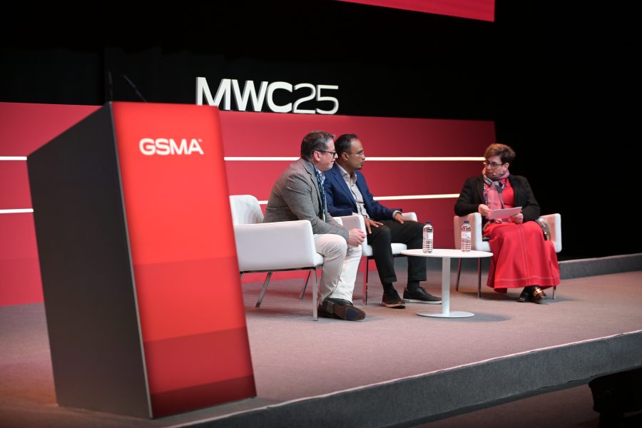 Three people sit on white chairs on a stage with a red backdrop, participating in a panel discussion at MWC25. A man on the left wears a gray blazer, the center man wears a dark suit, and a woman on the right wears a red outfit and scarf. A red podium with "GSMA" stands to the side.