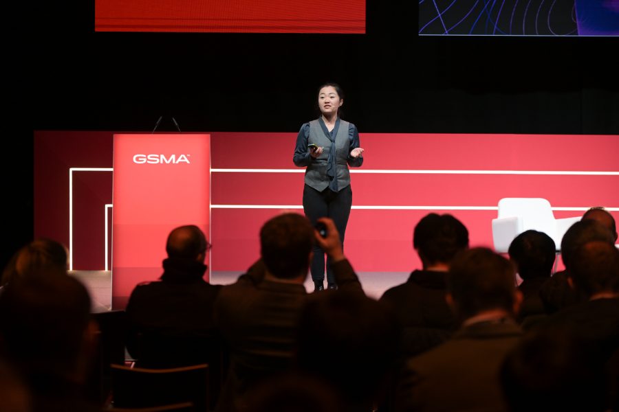 A person is speaking on stage at the GSMA event, standing in front of a red podium. The audience is seated, facing the speaker. The backdrop features red panels with white lines. A white chair is on the right side of the stage. The atmosphere is formal and engaging.