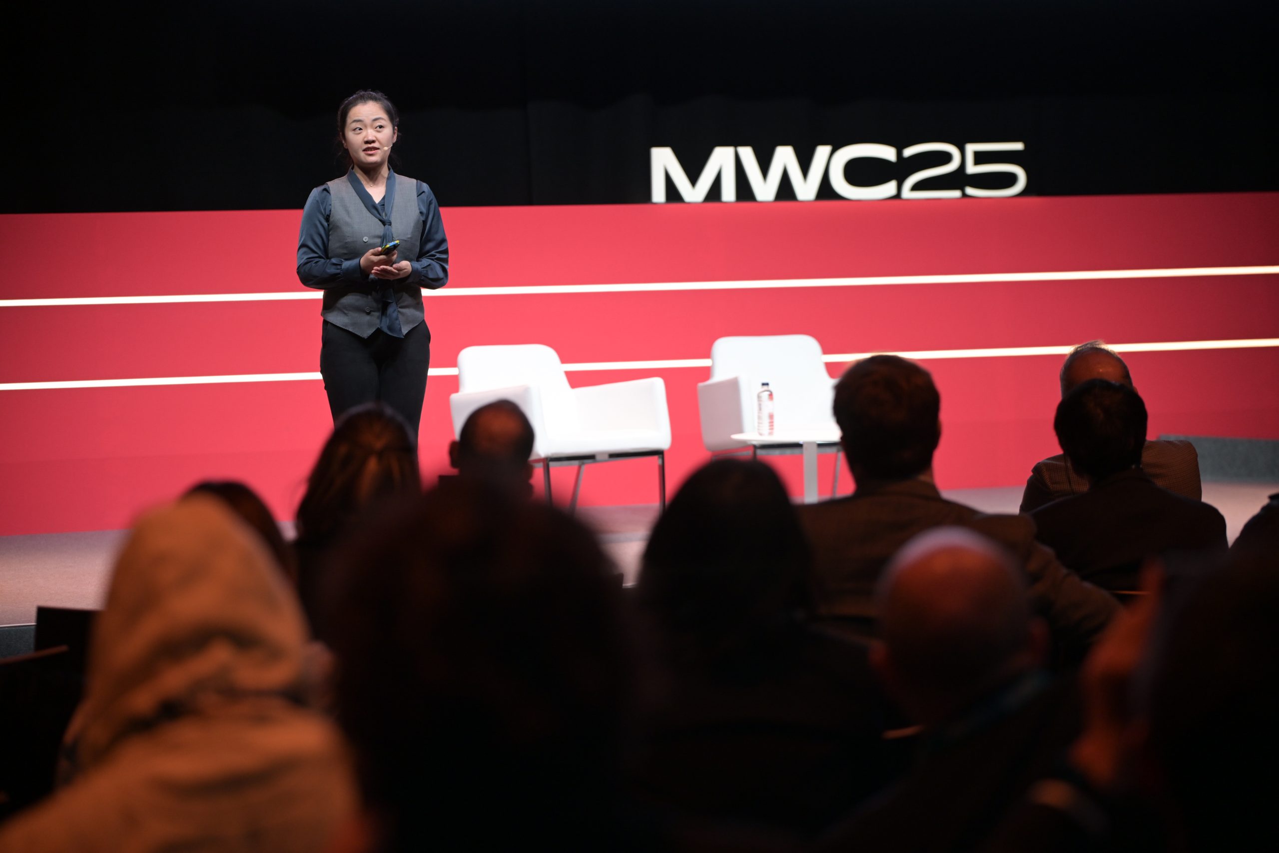A person stands on a stage delivering a presentation at MWC25. They're dressed in business attire. Behind them, a red backdrop features the MWC25 logo. Two white chairs and a small table are on stage. Several audience members are sitting, facing the speaker.