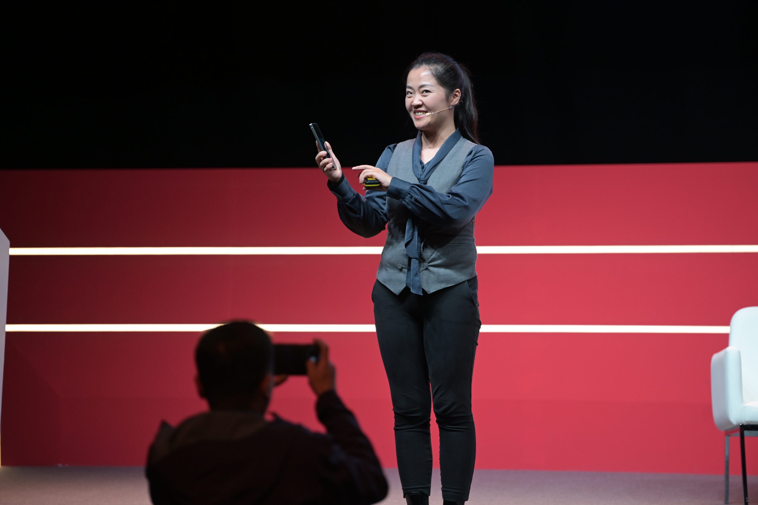 A person stands on stage holding a phone, smiling and gesturing, in front of a red backdrop with horizontal white lines. They are wearing a dark vest and pants. Another person in the foreground is capturing the moment with a phone. A white chair is partially visible on the right.