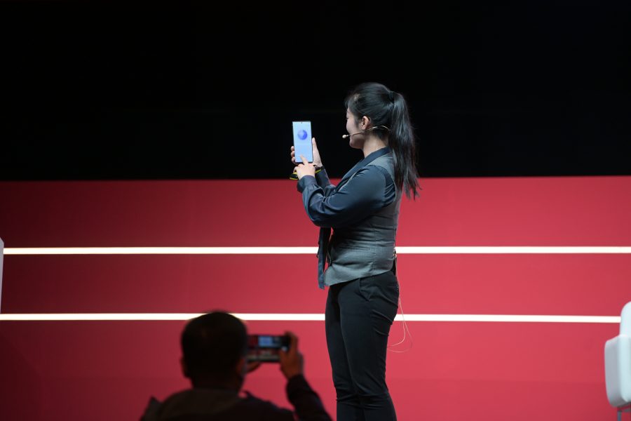 A person on stage holds up a smartphone, facing the audience. They are wearing a gray vest, black pants, and a microphone headset. The background is red with horizontal white lines. An audience member in the foreground takes a photo with another device.