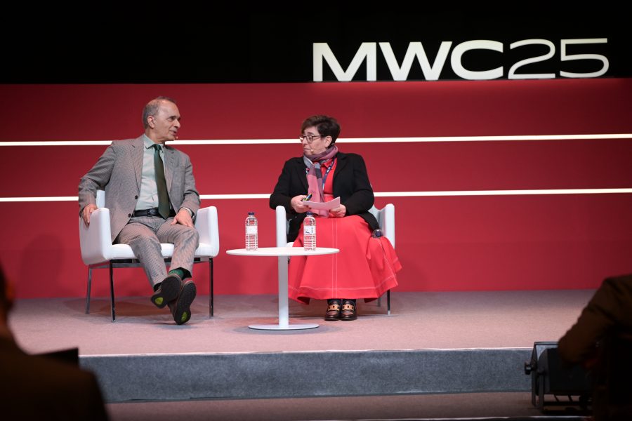 Two people sit on a stage at MWC25. The person on the left wears a gray suit, green tie, and dark shoes. The person on the right wears glasses, a dark jacket, and a red scarf and skirt. A small white table with water bottles is between them. They appear engaged in conversation.