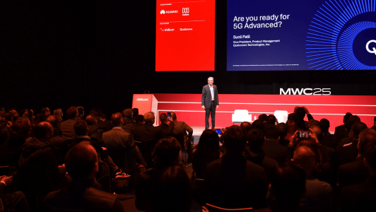 A speaker stands on stage at a conference with a presentation titled "Are you ready for 5G Advanced?" in the background. The audience is seated facing the stage. The event is branded with logos of Huawei, Vodafone, and Qualcomm. The stage backdrop is red and blue with "MWC25" displayed.