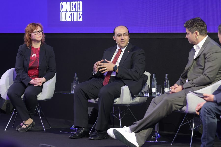 Three people are seated on stage, engaging in a panel discussion. A woman in a black blazer listens attentively, while a man in a suit and red tie speaks animatedly. A second man, in a gray suit, listens intently. The backdrop reads "Connected Industries." Bottles of water are nearby.