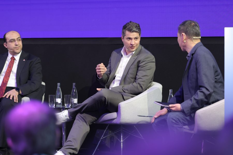 Three men are seated on stage in a panel discussion. The man in the middle, wearing a gray suit, gestures while speaking. The men on either side listen attentively. Two water bottles sit on the table between them. The background features a large purple screen.